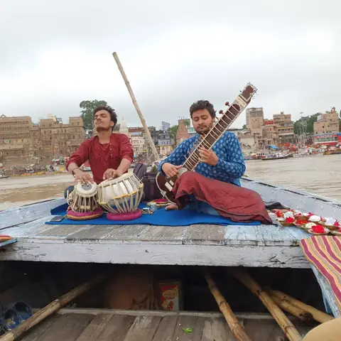 Dos músicos tocan el sitar y la tabla en la proa de una barca al amanecer en el Ganges, con los ghats de Varanasi al fondo
