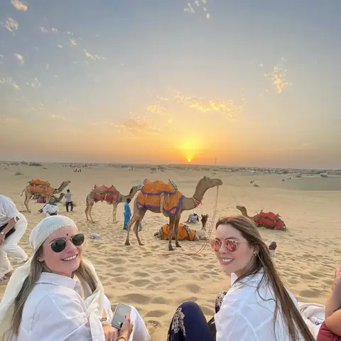 Dos viajeras sentadas en la arena contemplan la puesta de sol junto a los camellos en el desierto de Rajastán
