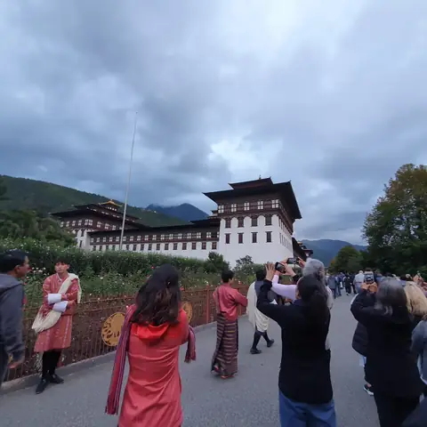 Viajeros y locales en traje tradicional caminan hacia un dzong bajo un cielo nublado en Bután